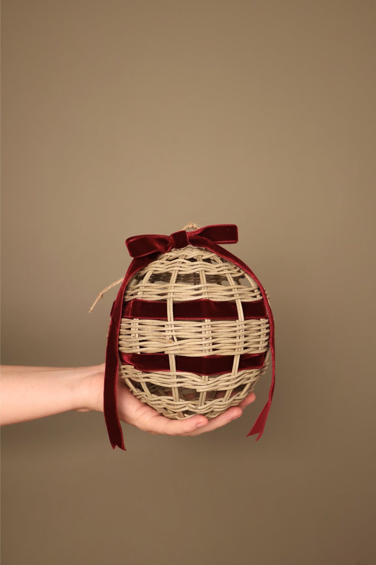Noël rattan bauble with a bordeaux velvet ribbon bow, held in a hand