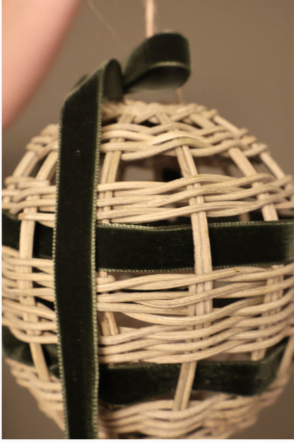 Close-up of a Noël rattan bauble with forest green velvet ribbon threaded through the weave and a long ribbon tail