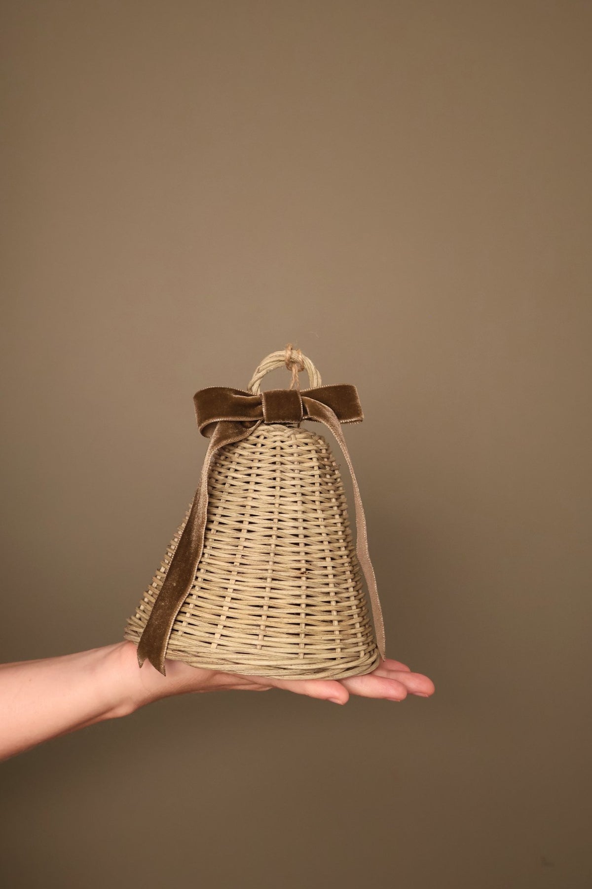 Medium Noël rattan bell with a chocolate velvet ribbon bow and jute loop, held in a hand