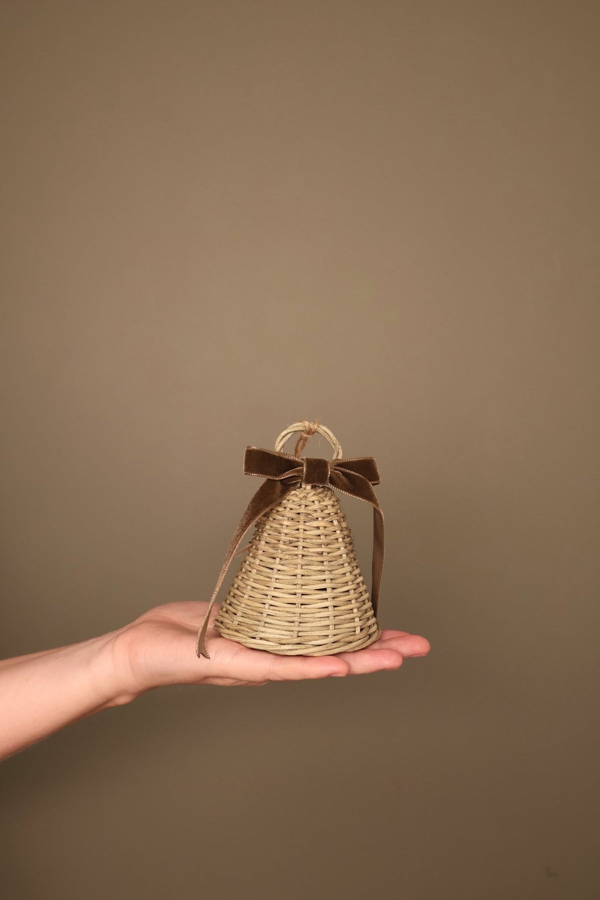 Small Noël rattan bell with a chocolate velvet ribbon bow and jute loop, held in a hand
